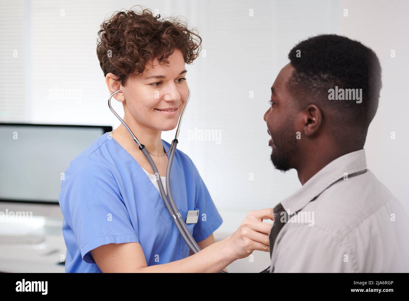 Horizontal medium close-up portrait of modern female doctor standing in front of male patient examining his heart beat and lungs using stethoscope Stock Photo