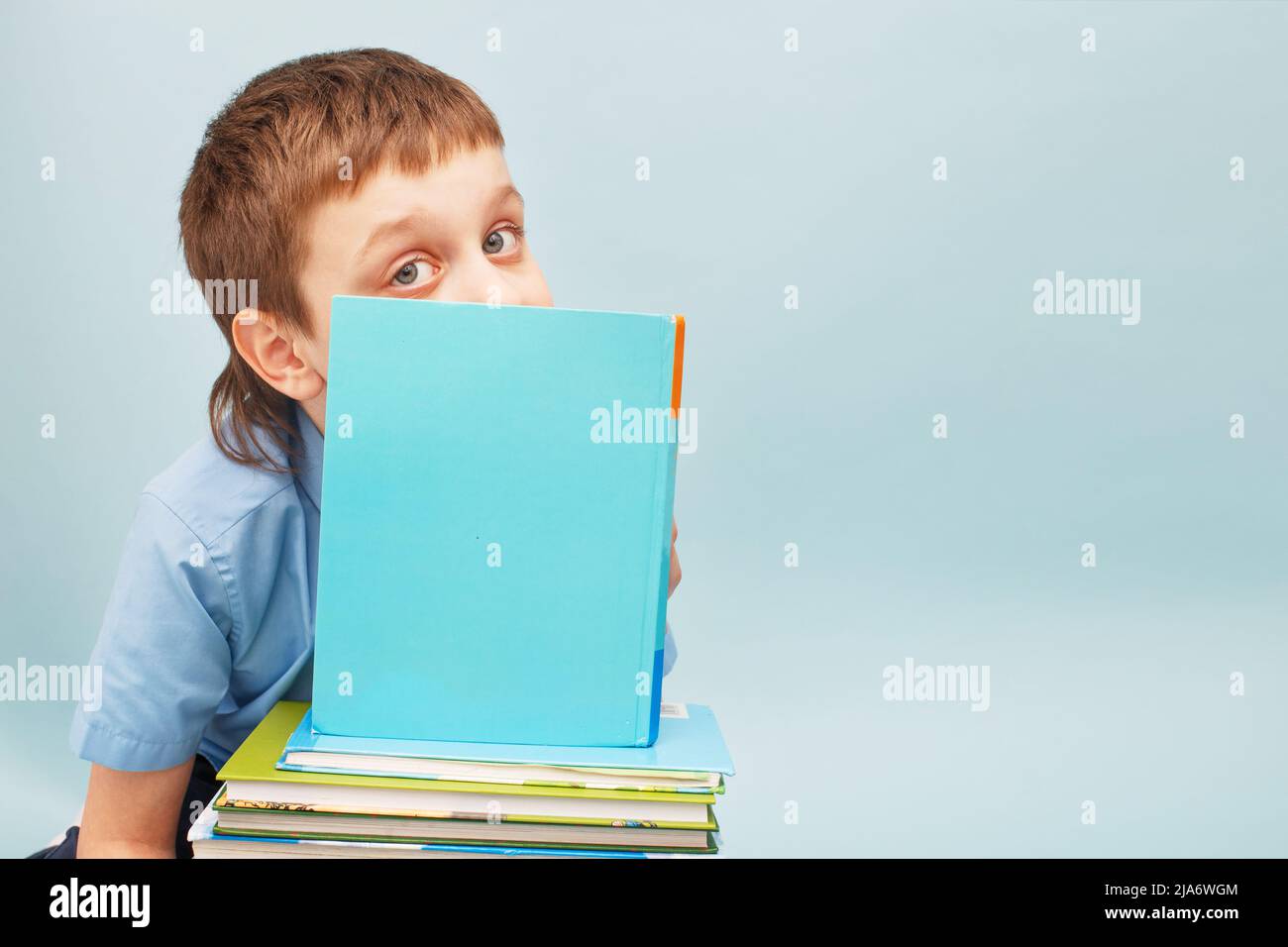 Schoolboy is sitting with a stack of books and reading and covers his face with a book isolated on a blue background Stock Photo