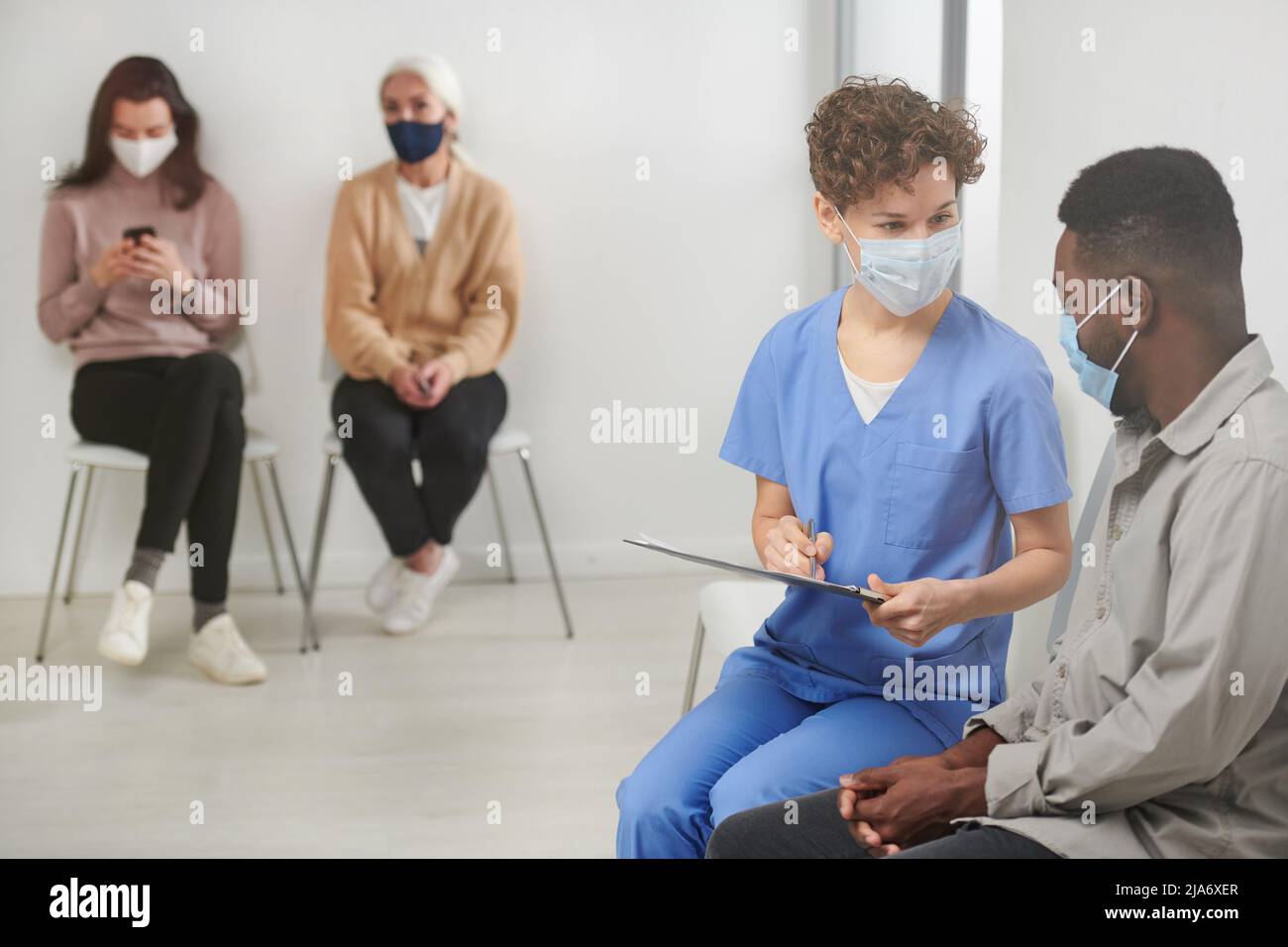 Horizontal shot of unrecognizable medical worker wearing mask sitting on chair in corridor next to patient asking him questions and making notes Stock Photo