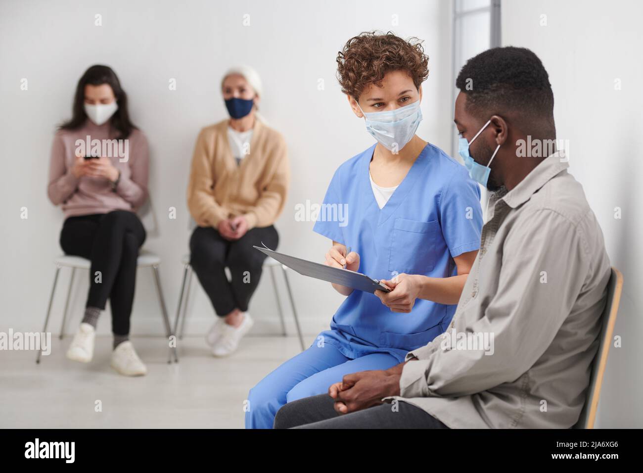 Horizontal shot of unrecognizable female medical worker wearing protective mask sitting on chair in corridor next to patient talking to him about test Stock Photo
