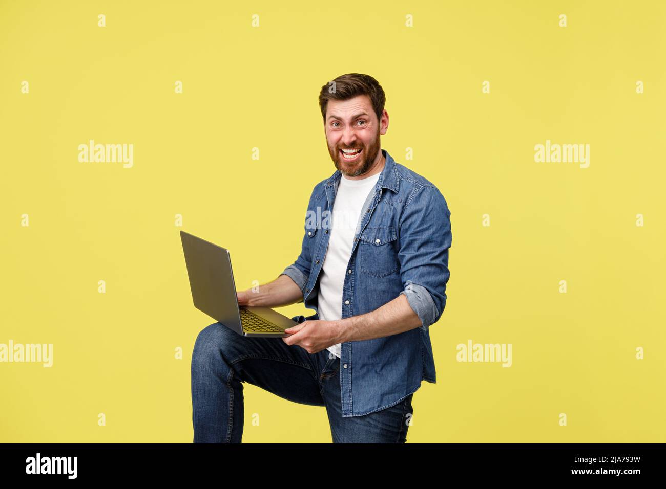 Crazy smiling man with beard holding laptop on leg and looking at camera. Studio shoot Stock Photo