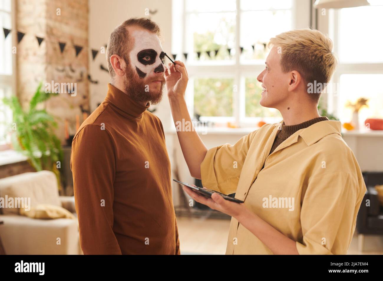 Smiling young blond-haired woman in yellow shirt applying makeup to man while preparing him for Halloween party Stock Photo