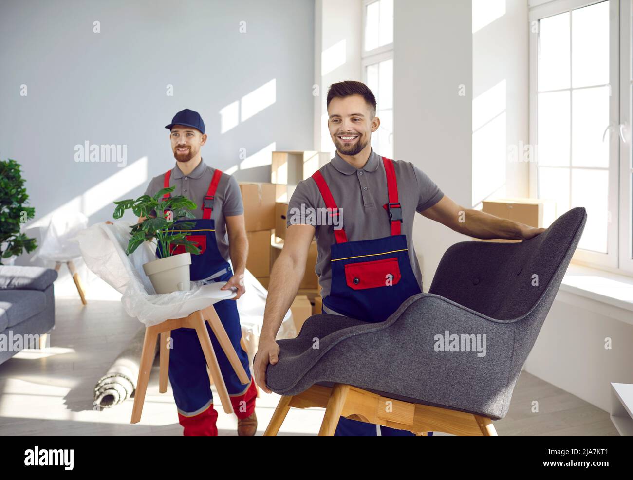 Two happy young moving company workers carrying chairs, armchairs and house plants Stock Photo