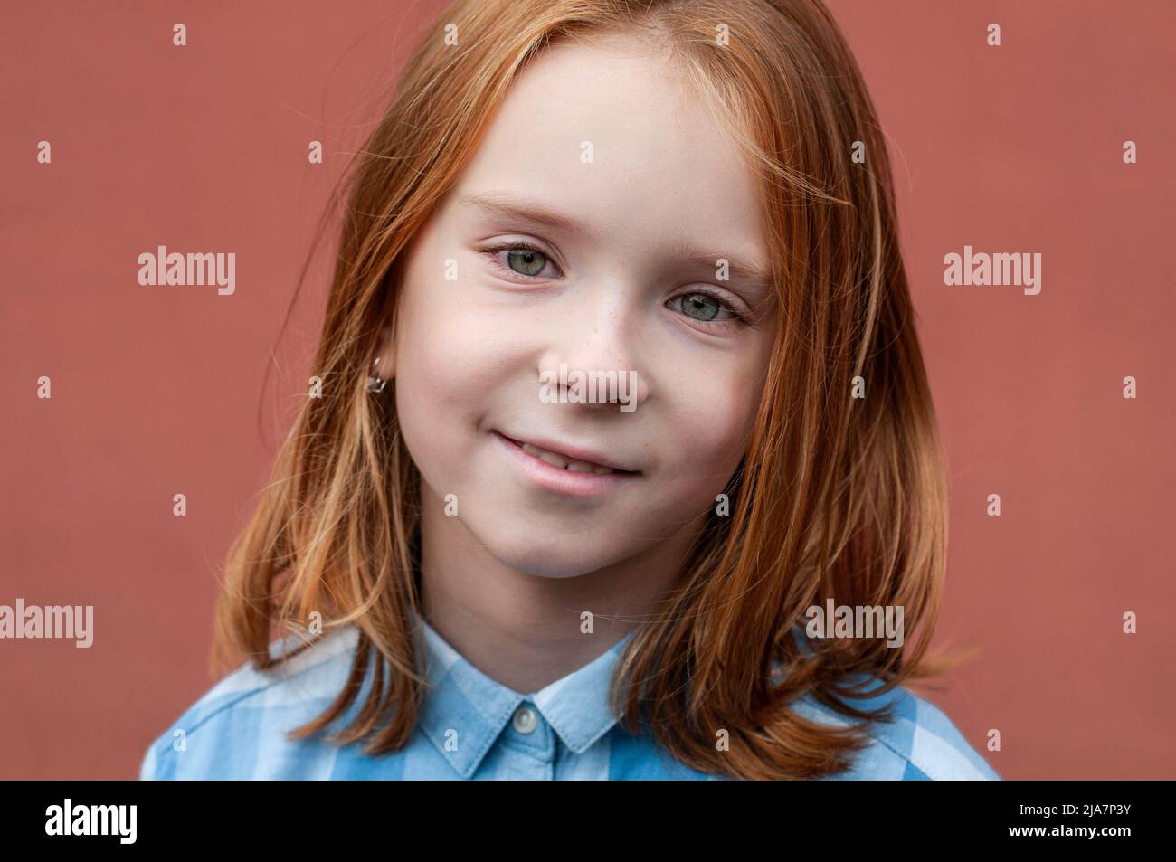 Portrait of a red-haired child with green eyes 8 years old in a blue shirt on a teracot background. Stock Photo