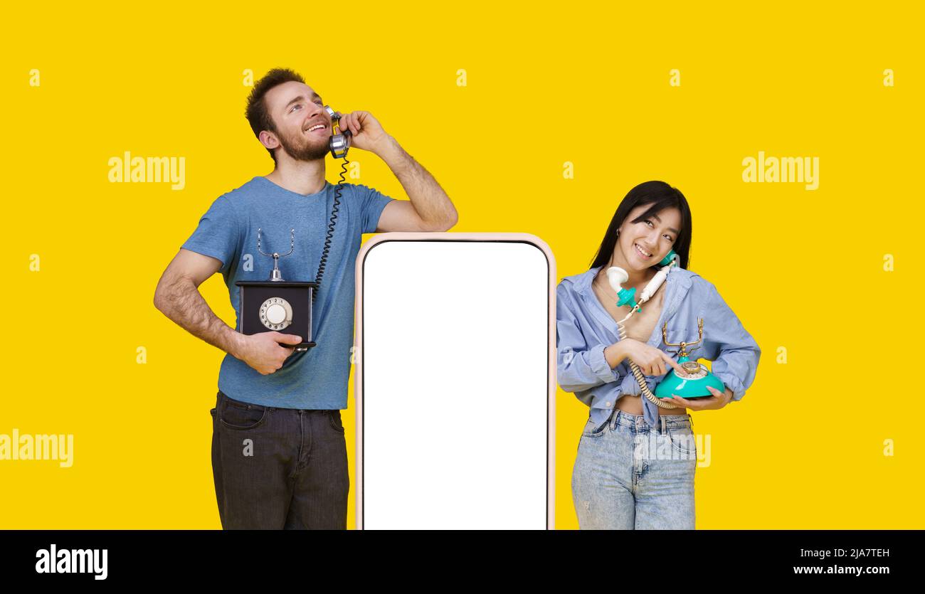 Asian girl and caucasian guy in love talking on vintage phones leaned on huge smartphone with blank screen, looking at each other isolated on yellow background. Product placement. Stock Photo