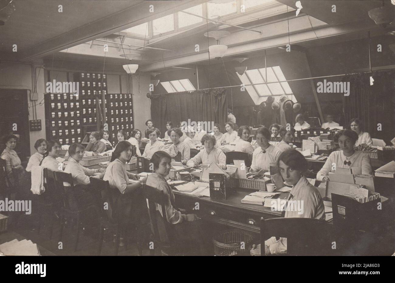 Women's Claims Department, National Health Insurance, Liverpool Victoria Approved Society (LVAS): women office workers seated at long tables. Filing cabinet draws of case notes and baskets containing papers are on the tables. Hats and coats are hung up at the back of the office, partially hidden by a curtain. Light is coming into the attic room from a skylight and slanting windows Stock Photo