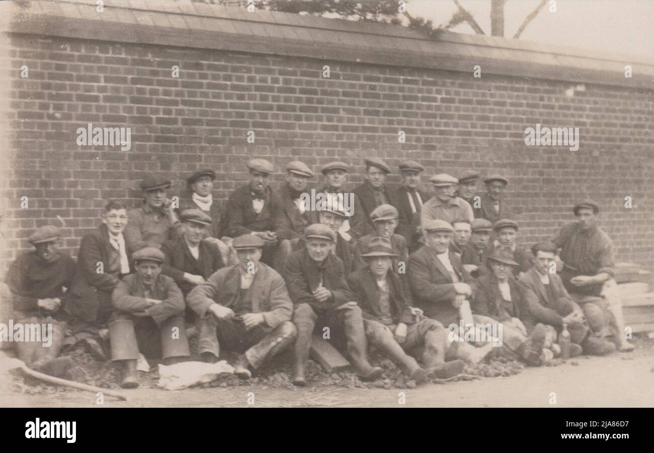 Group of navvies / manual labourers on a break, leaning against a brick wall. Almost all are wearing flat caps & some are smoking cigarettes or pipes. A shovel is on the ground on the left and several of the workers have bottles of drink Stock Photo