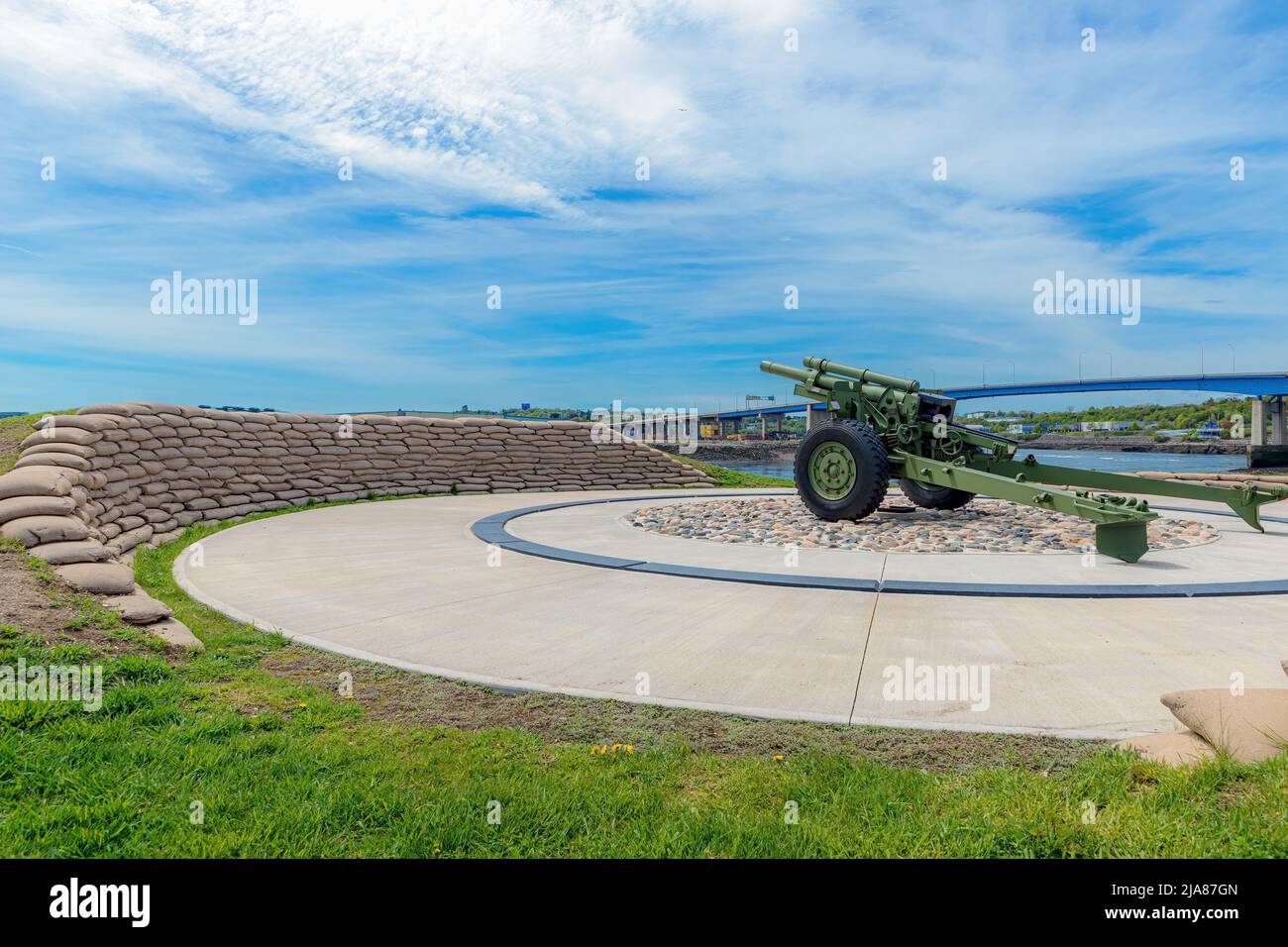 A 105mm howitzer in Saint John NB behind a curve of sandbags. Harbor bridge in background, blue sky with cloud above.Gun is on concrete and rock. Stock Photo