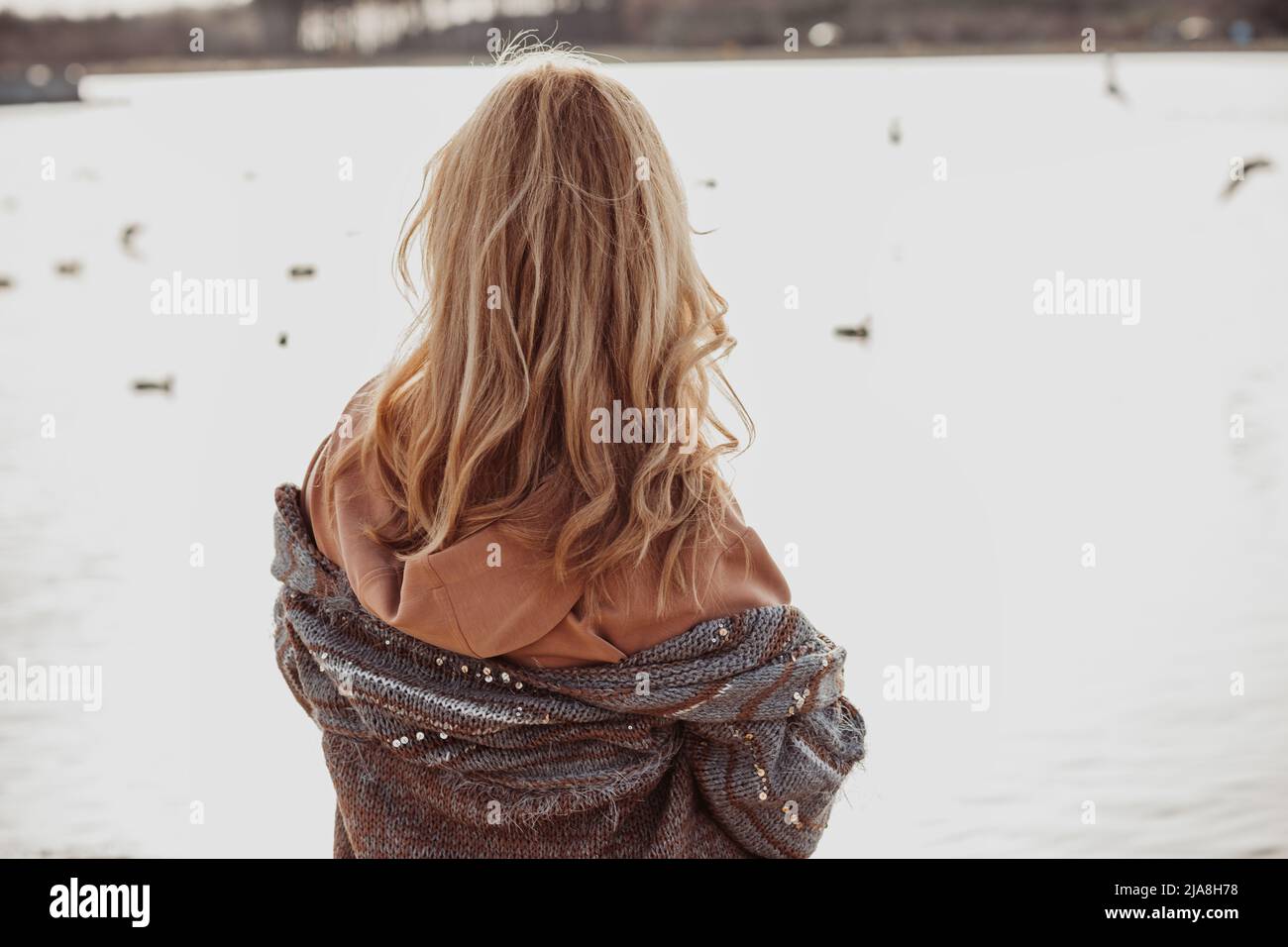 Close-up of woman with long wavy fair hair wearing brown sweatshirt, woolen cardigan, standing on sandy beach of river. Stock Photo