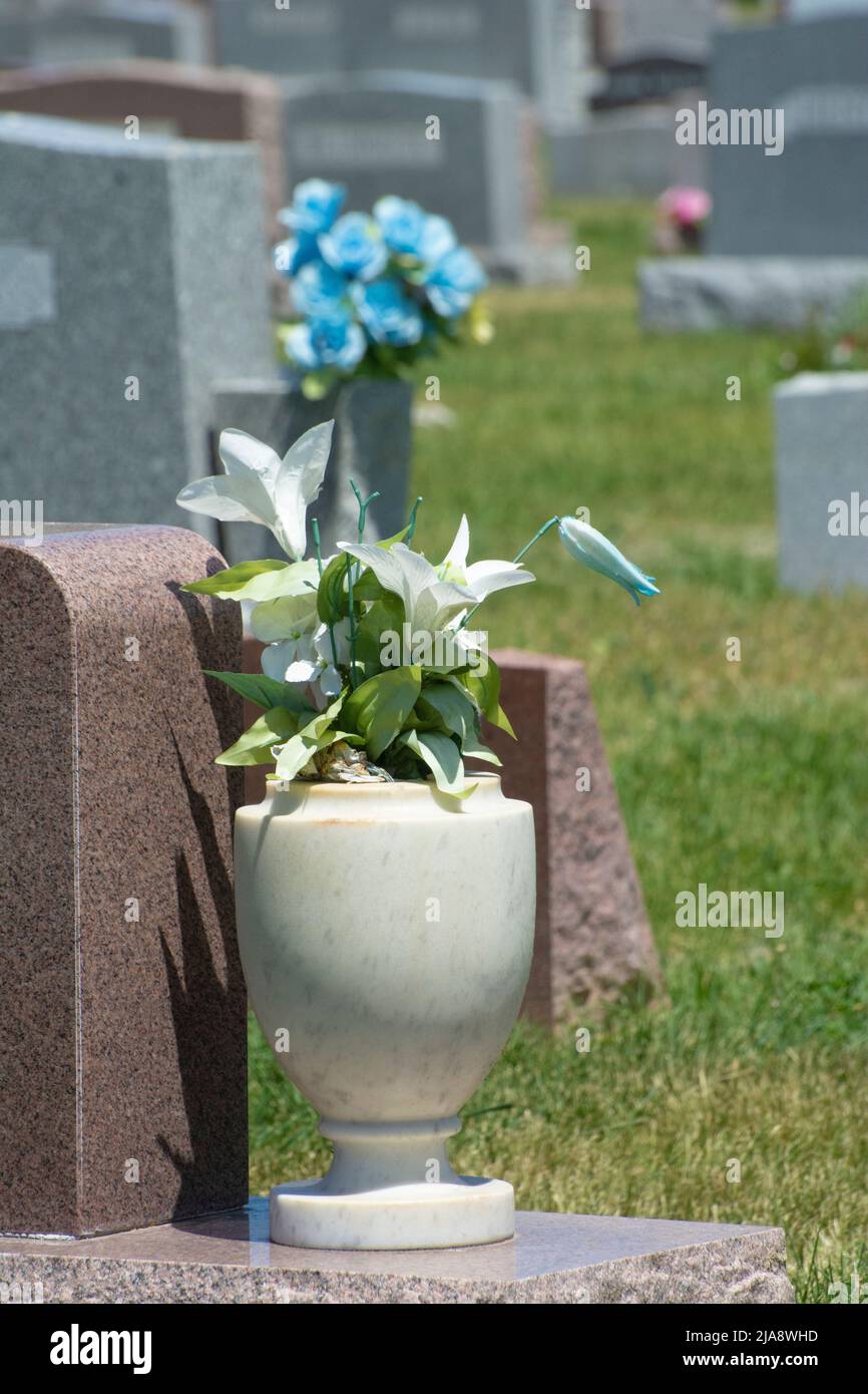 Granite vase of artificial all weather fabric lily flowers next to gravestone in cemetery Stock Photo