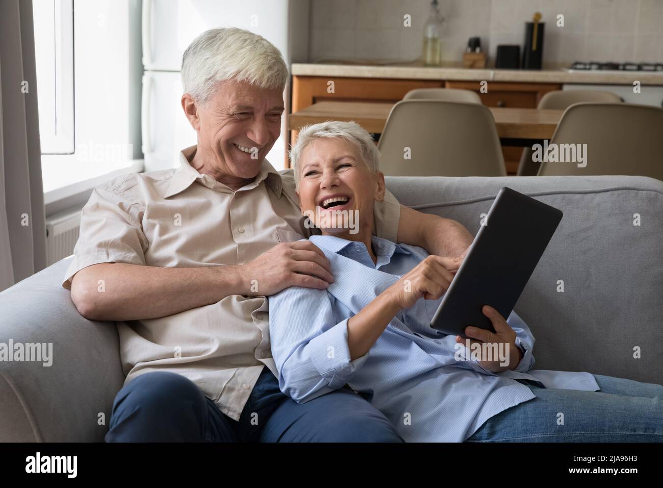 Excited older grandparents hug on couch laugh using digital touchpad Stock Photo