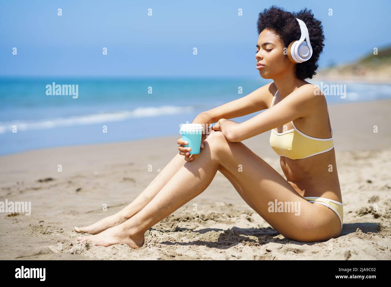 African American girl sitting on the beach with headphones, sipping a drink from a take-away cup. Stock Photo