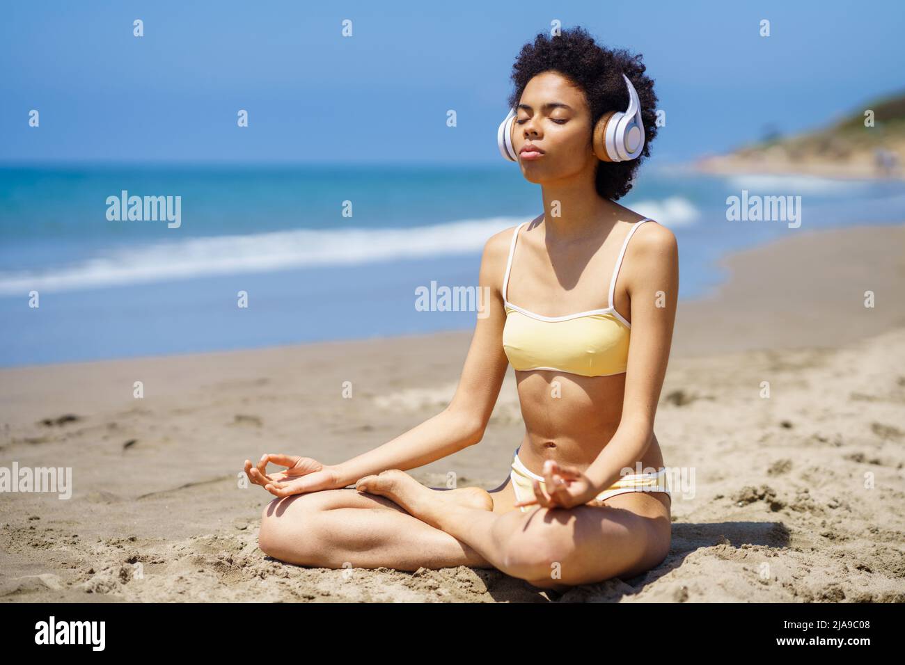 Black girl in lotus pose doing mindfulness listening to a meditation with wireless headphones. Stock Photo