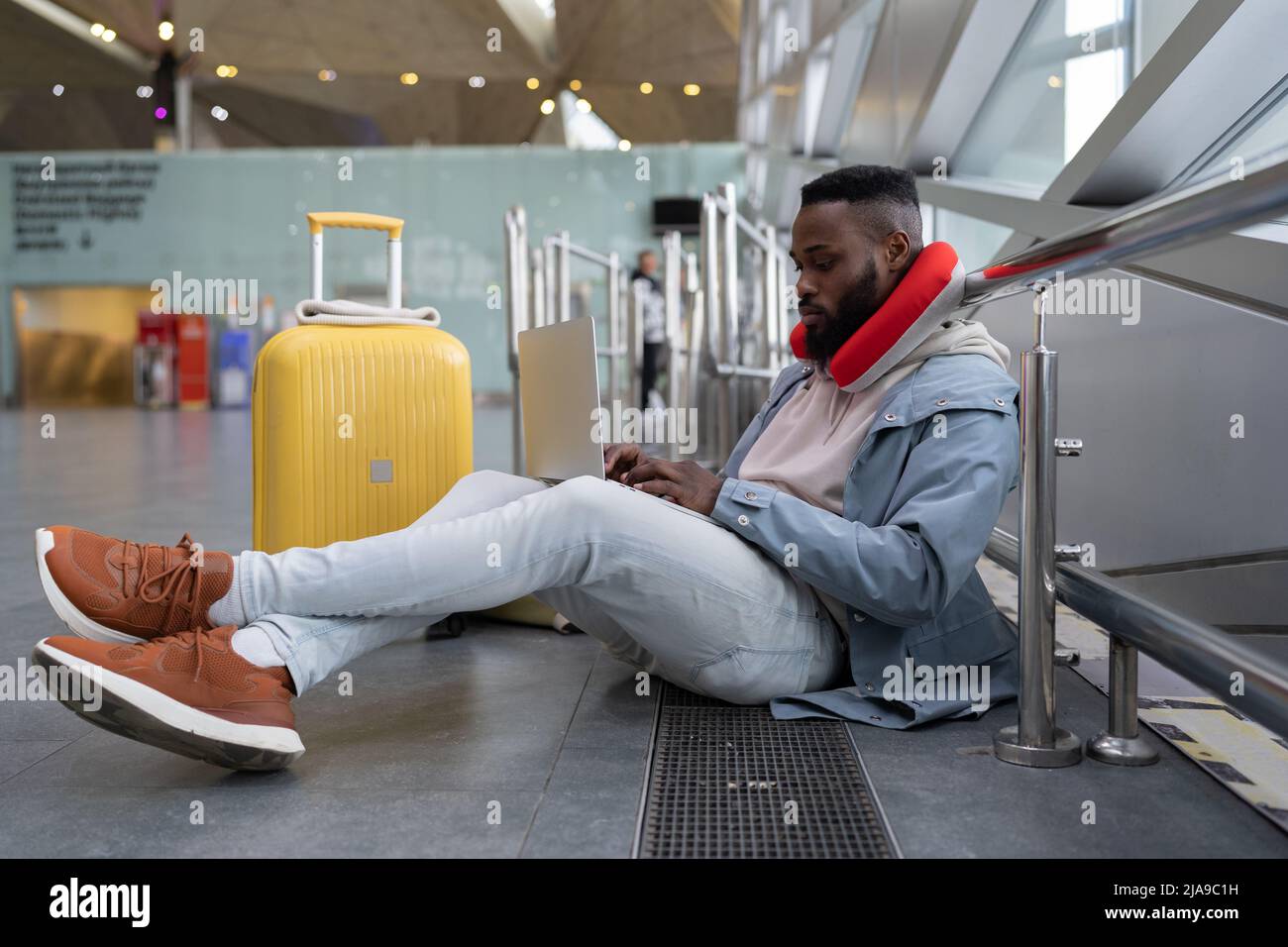 Pensive young black man sits cross-legged together waiting for flight using computer next to window. Stock Photo