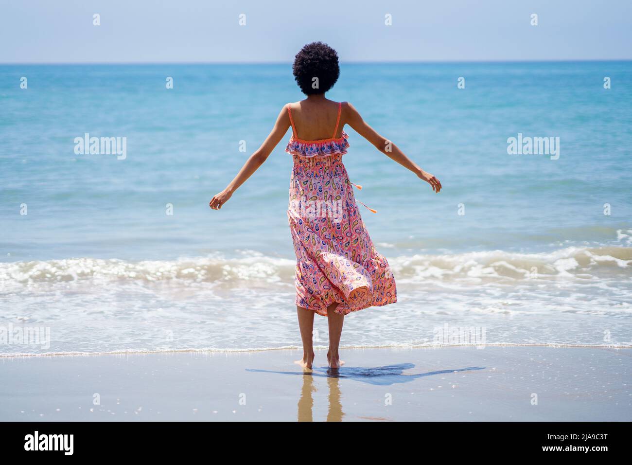 Rear view of black woman with afro hair in summer dress, opening her arms looking out to sea. Stock Photo