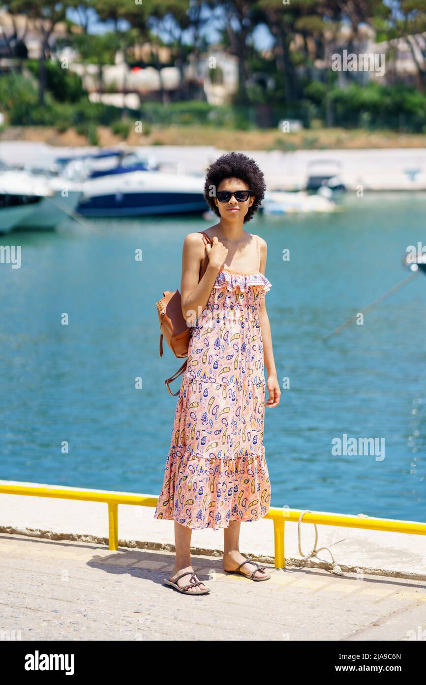 Happy black female with afro hairstyle in summer dress, walking through a harbour full of boats. Stock Photo