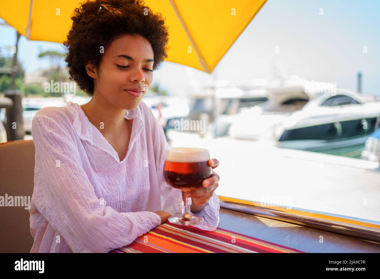 Black woman drinking a glass of cold beer with foam on the terrace of a bar in a coastal area. Stock Photo