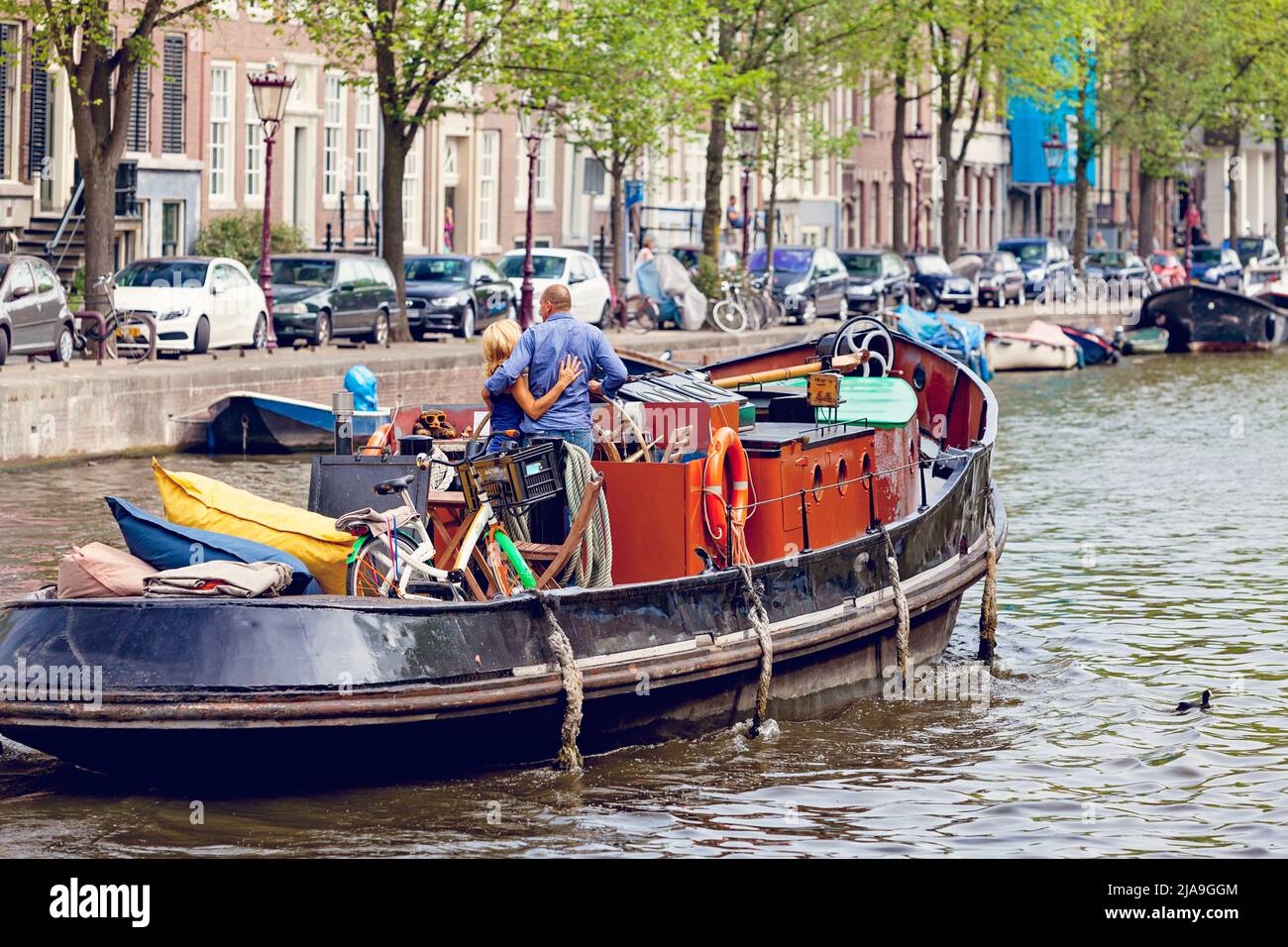 man and woman, couple, embrace travelling in a barge, boat, along a tree lined Amsterdam city canal. Stock Photo