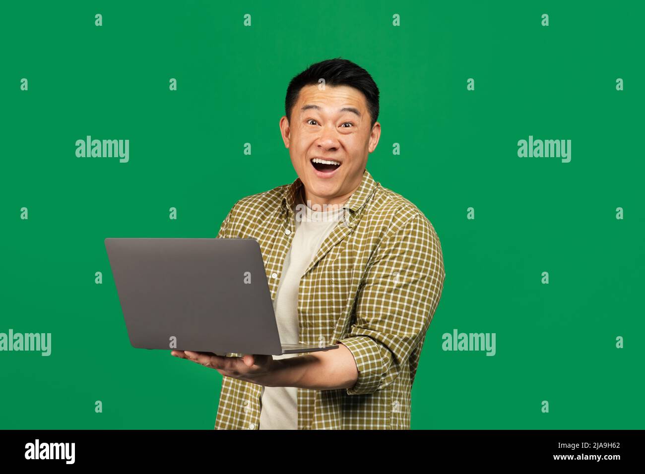 Excited asian mature man holding laptop and looking in surprise at camera, opening his mouth, green background Stock Photo