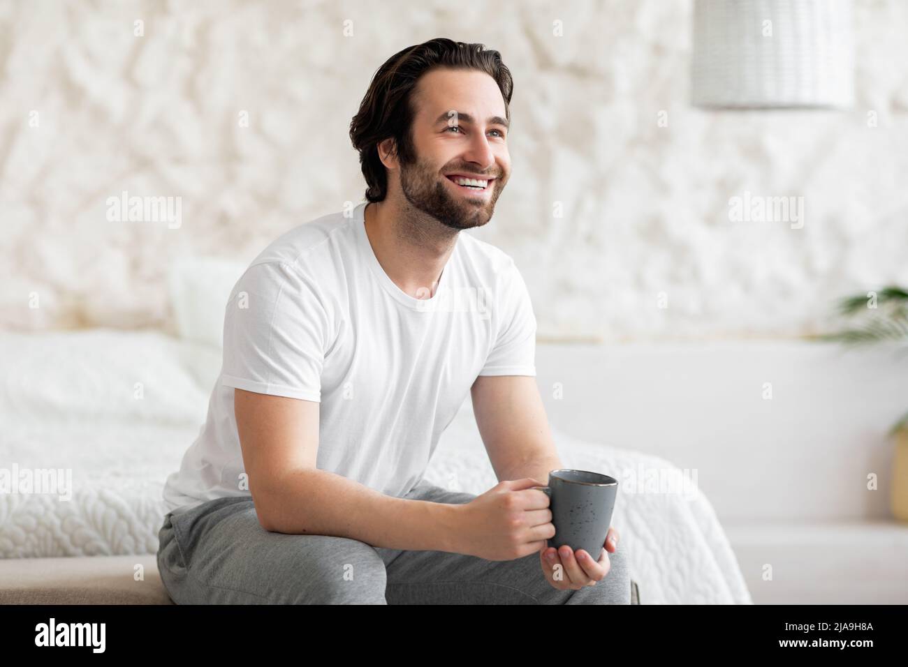 Happy bearded young man sitting on bed, drinking coffee Stock Photo