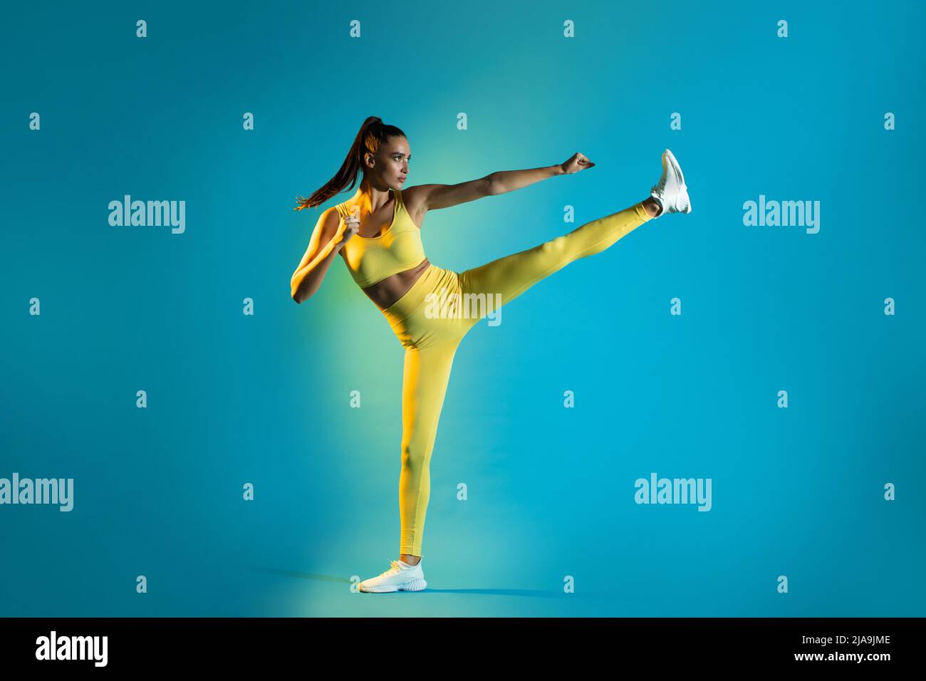 Athletic Female Doing Karate Side Kick Clenching Fists, Blue Background Stock Photo