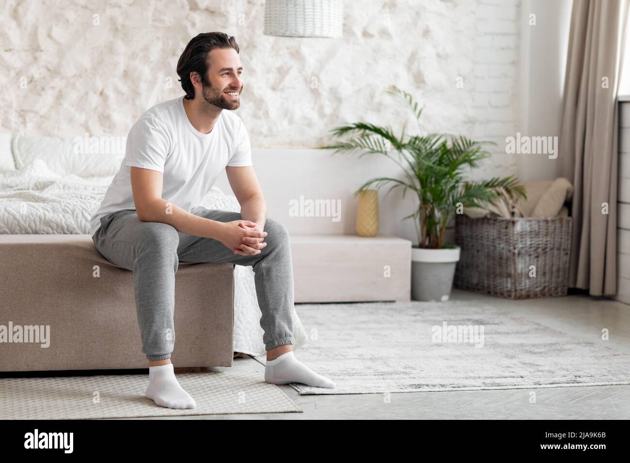 Happy bearded young man sitting on bed at home Stock Photo