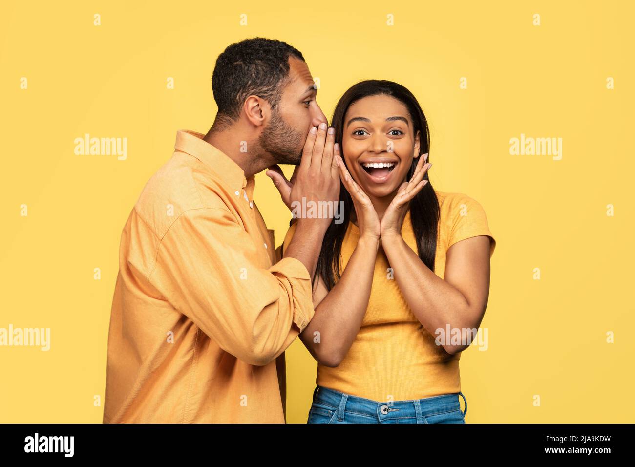 Young black guy sharing secret or whispering gossip into his girlfriend's ear on yellow studio background Stock Photo