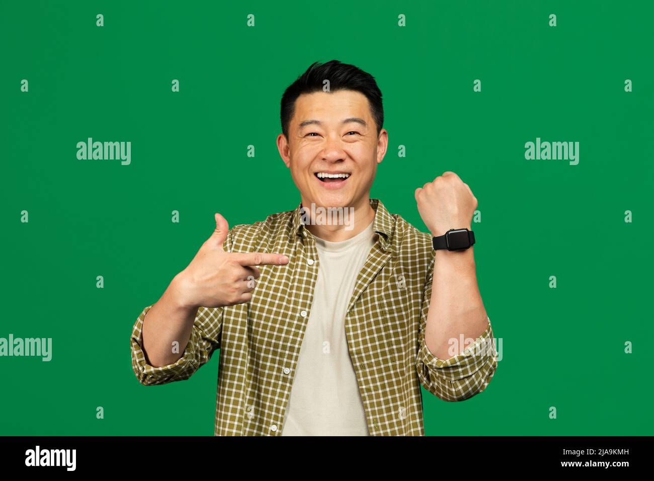 Happy asian mature man wearing and pointing at smartwatch, recommending modern gadget, posing over green background Stock Photo