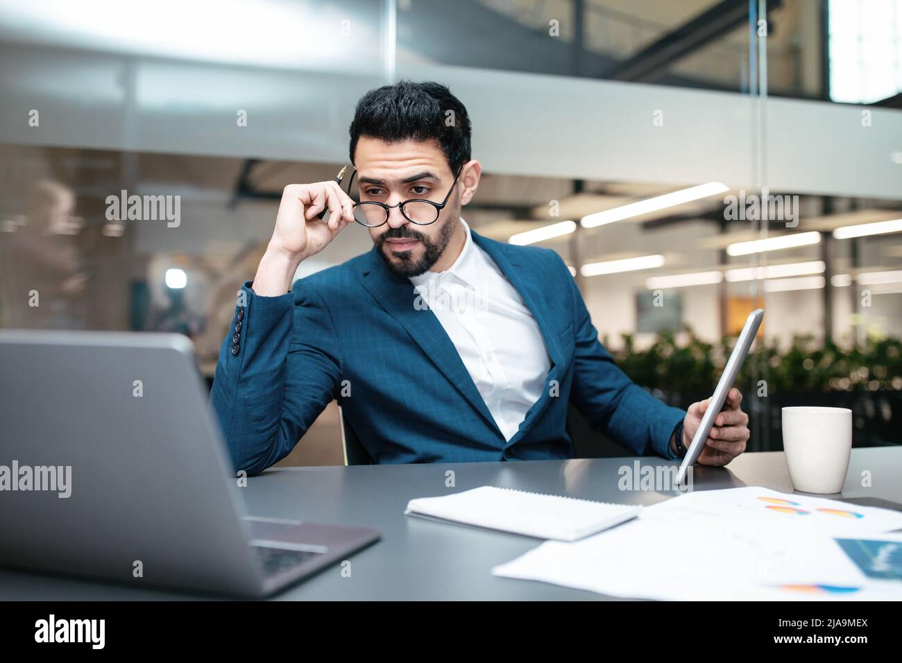 Serious busy attractive millennial arab businessman with beard in glasses works with computer and documents Stock Photo
