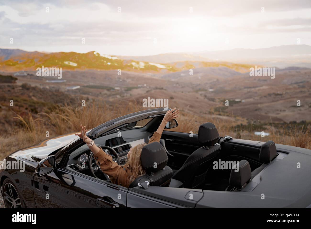 Rear of young pretty happy female holding hand up feeling free sitting in convertible car Stock Photo