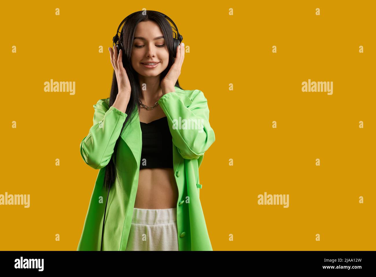 Happy brunette girl with black earphone listening music inside. Portrait view of female music fan in green jacket with closed eyes, enjoying sound, isolated on orange background. Concept of music fan. Stock Photo