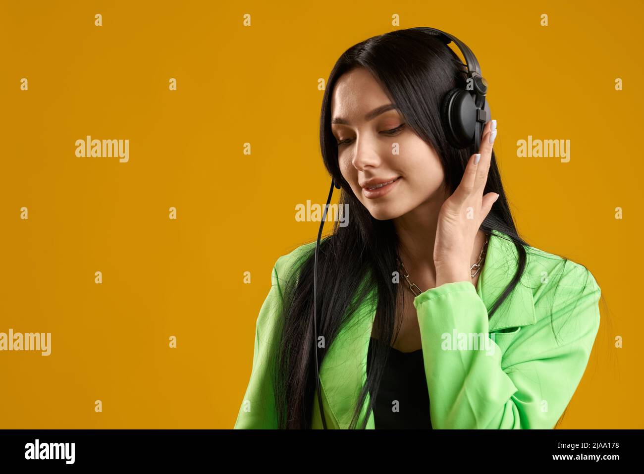 Young cheerful caucasian female with headphones, listening music indoors. Delighted female meloman in trendy blazer enjoying tune, isolated on yellow studio background. Concept of music.  Stock Photo