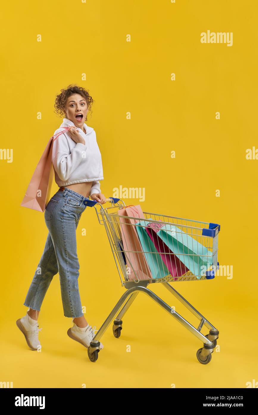 Shocked woman carrying shopping cart, with bright paper bags inside. Side view of surprised girl with shopping trolley, standing on tiptoe, isolated on orange studio background. Concept of shopping.  Stock Photo