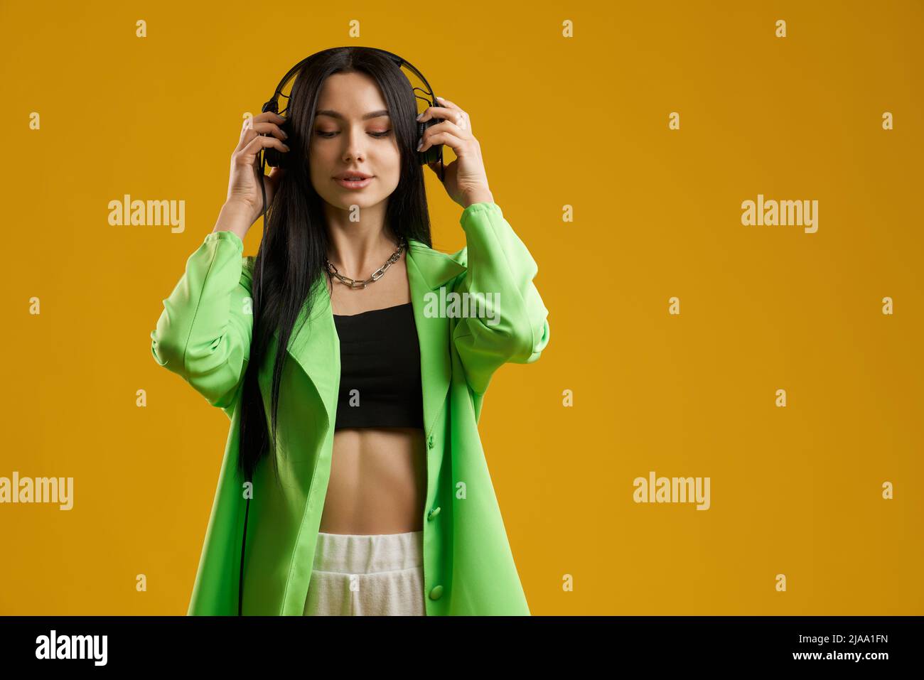 Mindful female in stylish jacket and crop top wearinging headphones on head outdoor. Portrait view of inspired brunette lady listening music with closed eyes, isolated on yellow. Concept of meloman. Stock Photo