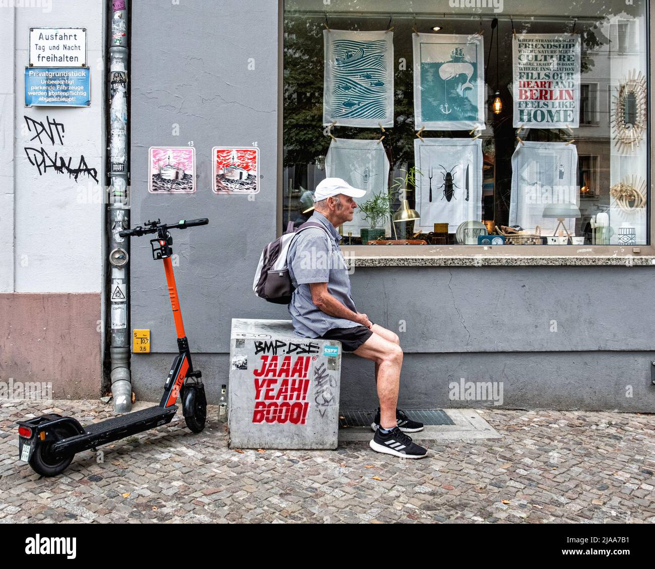 Elderly, senior man sitting outside a shop display window next to a Circ e-scooter In Kastanienallee, Prenzlauer Berg, Berlin Stock Photo