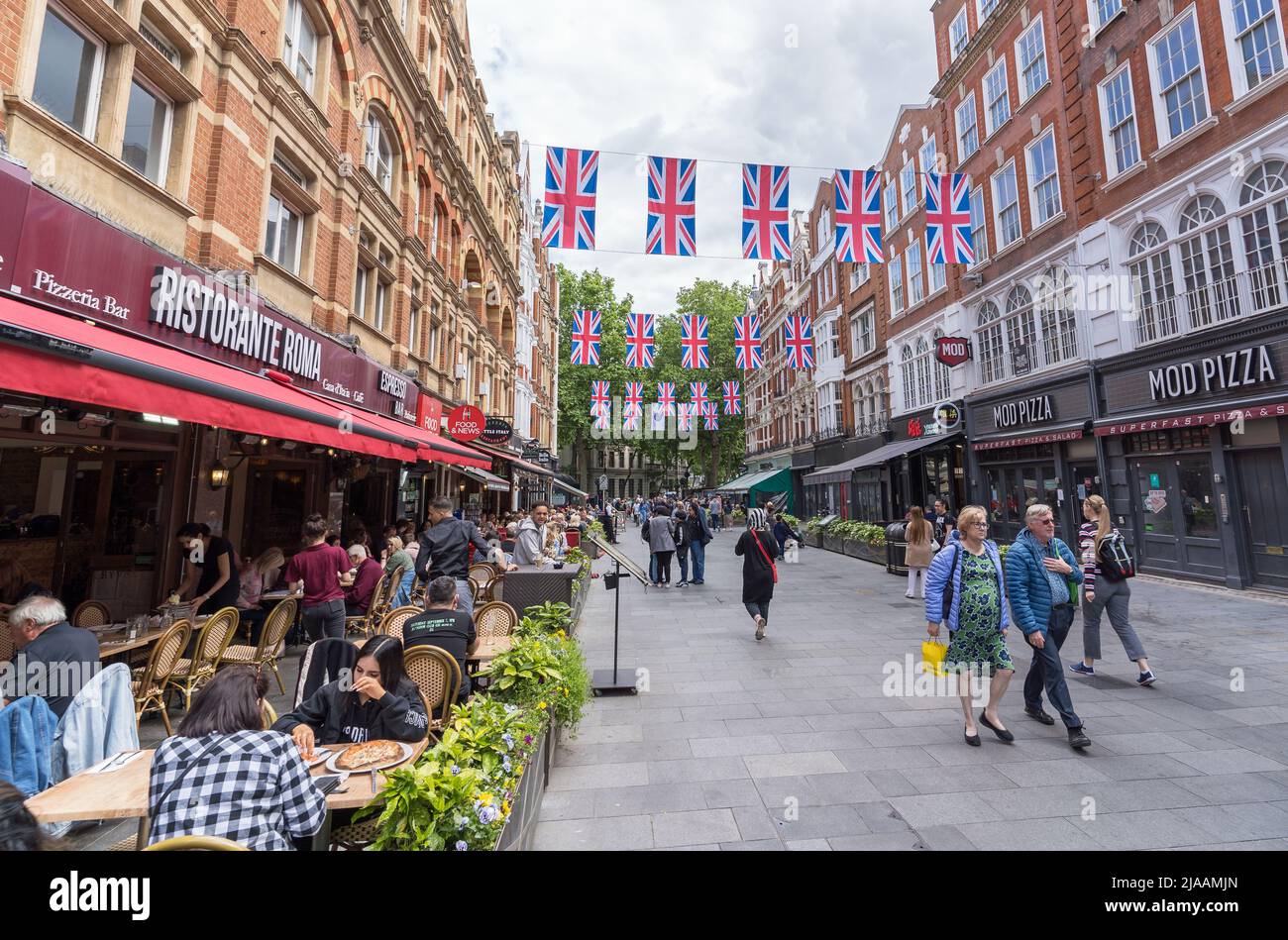 People dining outside in restaurants and cafes in Leicester Square with union Jack flags hanging over the street. London Stock Photo