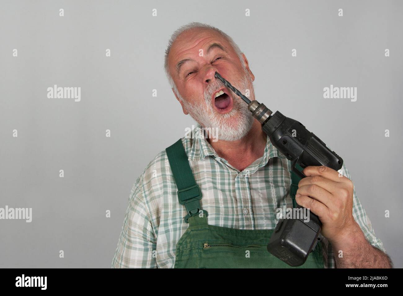 Taken literally in the nose picking. An old craftsman picks in his nose with his drilling machine, in front of a white wall. Stock Photo
