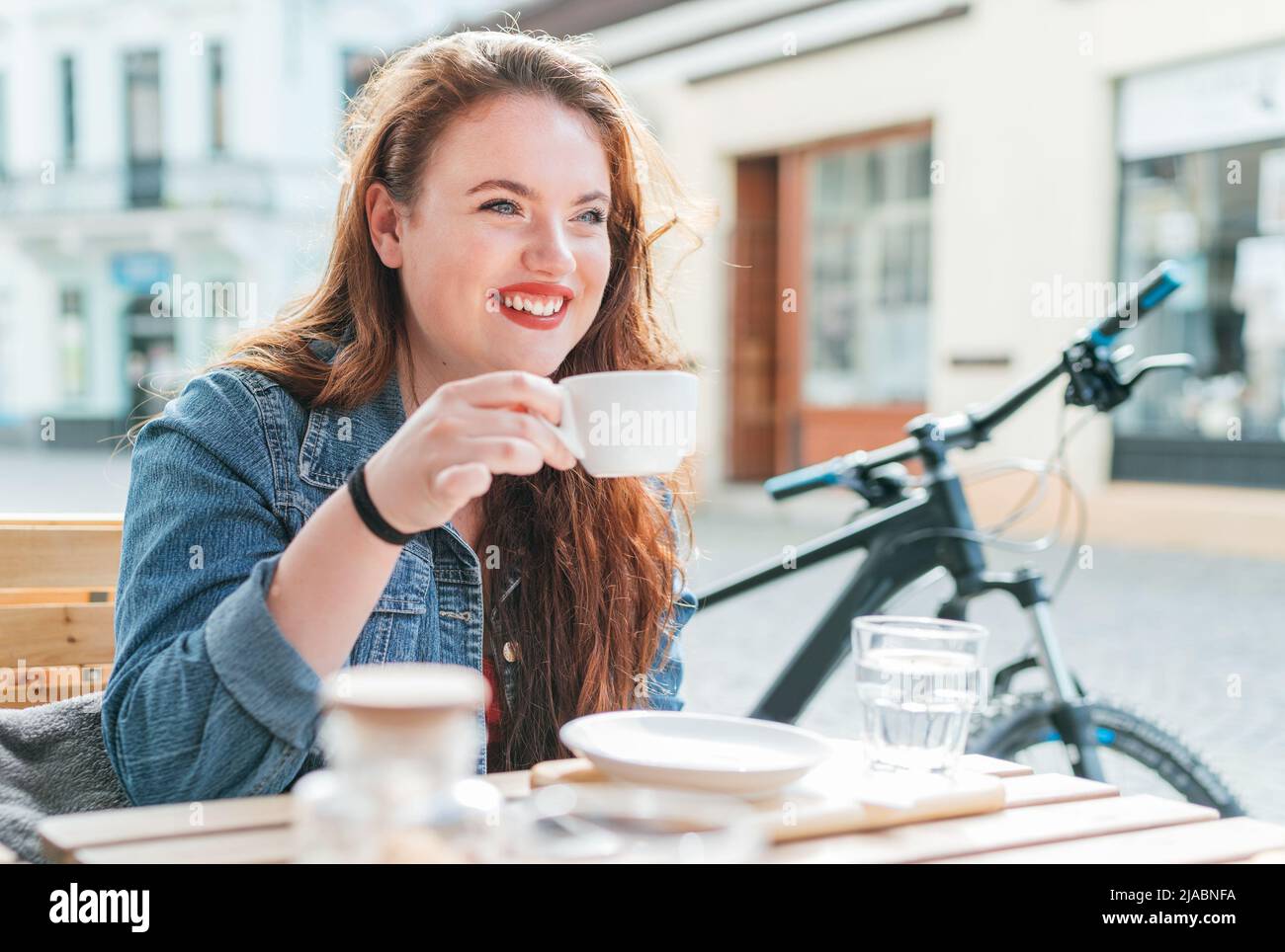 Portrait of sad red curled long hair caucasian teen girl sitting on a cozy cafe outdoor terrace on the street and drinking coffee. Young woman taking Stock Photo