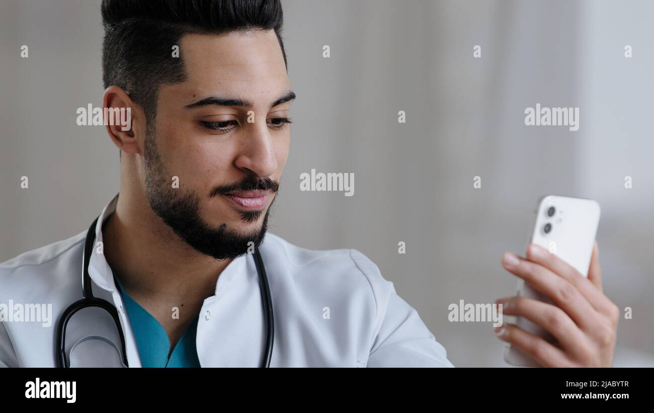 Confident qualified physician male arabian doctor use smartphone social medical app consult clients online in clinic cabinet remote communication give Stock Photo