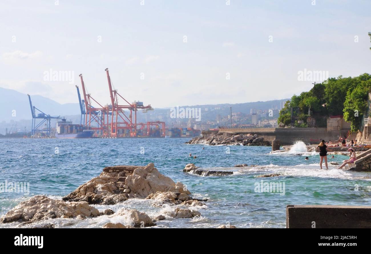 Cranes Loading Cargo Containers at Adriatic Gate Port in Rijeka.Pecine beach, people swimm in Adriatic Sea. Port of Rijeka. Gantry Cranes,Cargo Termin Stock Photo