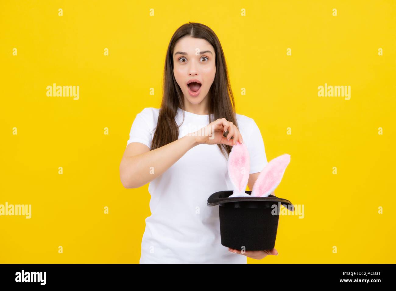 Happy Easter. Portrait of young excited woman with bunny ears isolated on yellow studio background. Rabbit ears appear from the magic hat. Stock Photo