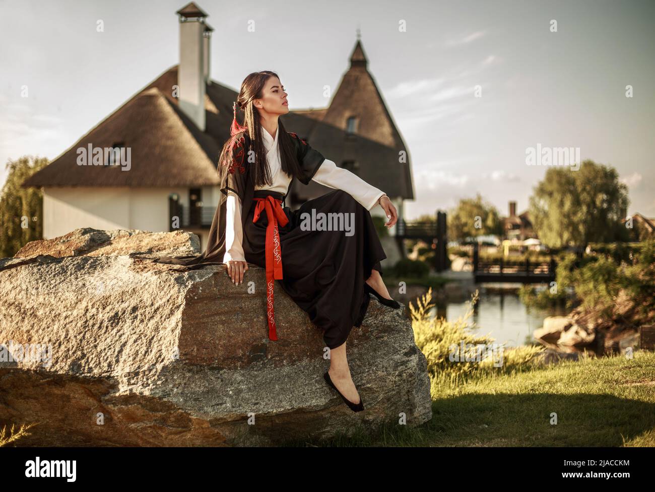 Pensive asian girl looking with hope sitting on a stone in a traditional kimono in the garden on the background of an ancient settlement and sky Stock Photo