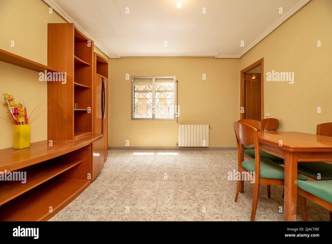 Living room with cherry-colored square folding dining table against one wall, light stoneware floor and cream-colored walls and matching bookcase on t Stock Photo