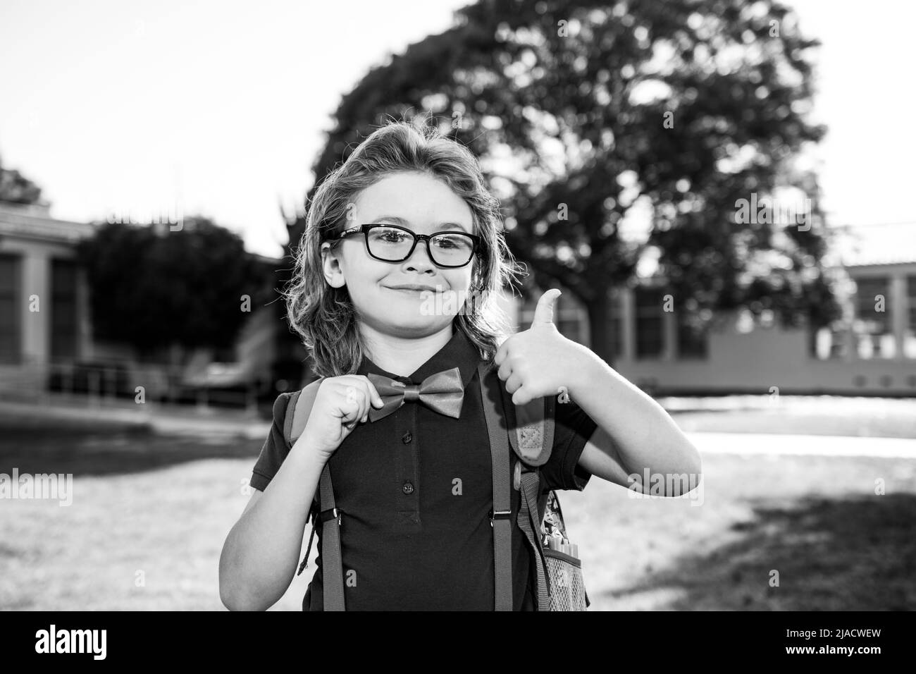 Happy smiling schoolboy pupil in glasses with thumb up is going to school. Child with school bag. Kid outdoors of the school. Stock Photo