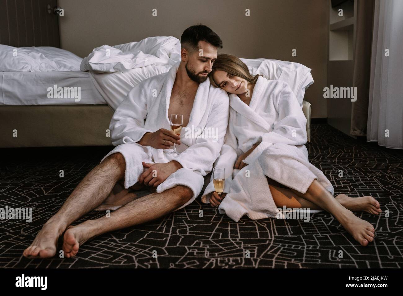 Happy young couple sitting on the floor near the bed in hotel with glasses of champagne Stock Photo