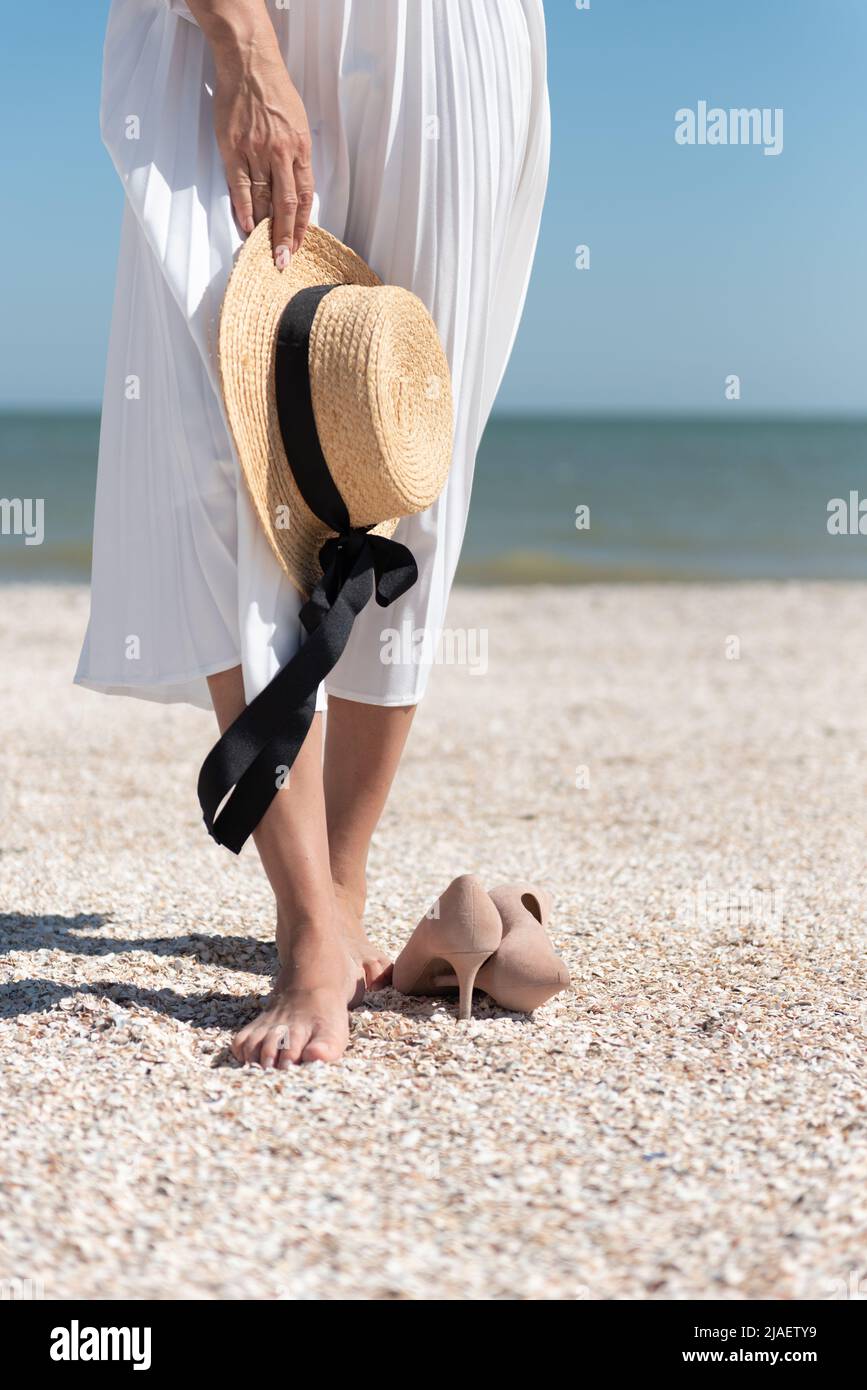 A barefoot woman in a white skirt with a straw hat in hand stands on the seashore, her beige high-heeled shoes lie on the sand. Stock Photo