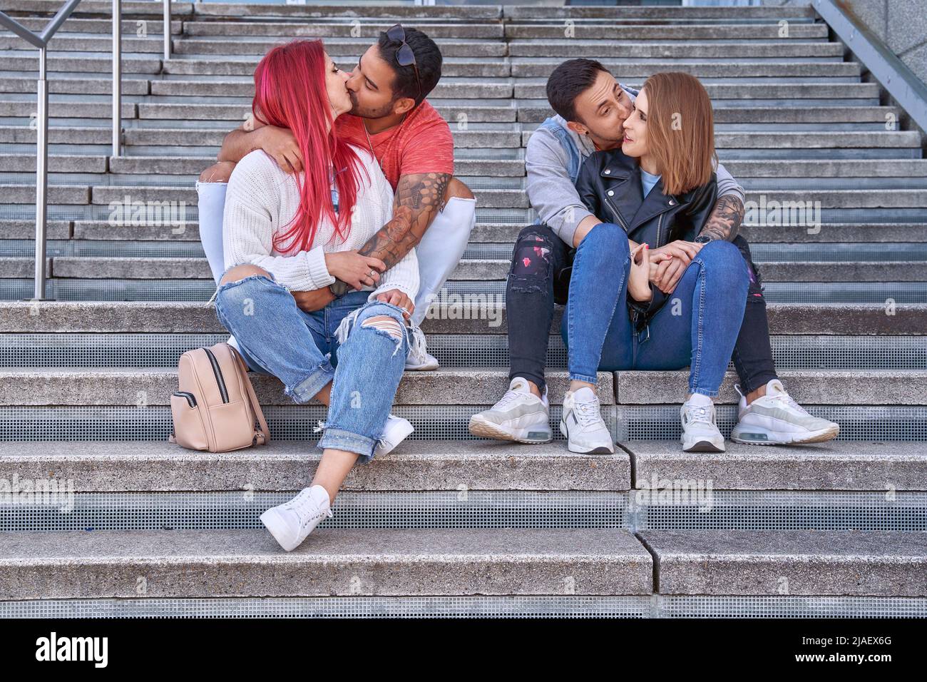 Two young couples kissing and hugging while enjoying time together sitting on stairs outdoors. Relationship and love concept. Stock Photo