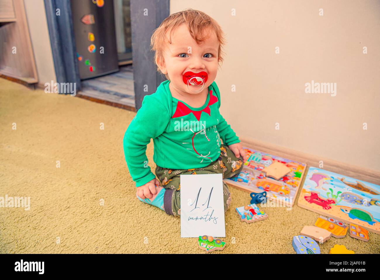 Cheerful baby with a pacifier in his mouth and a 11 months old milestone card with his toys