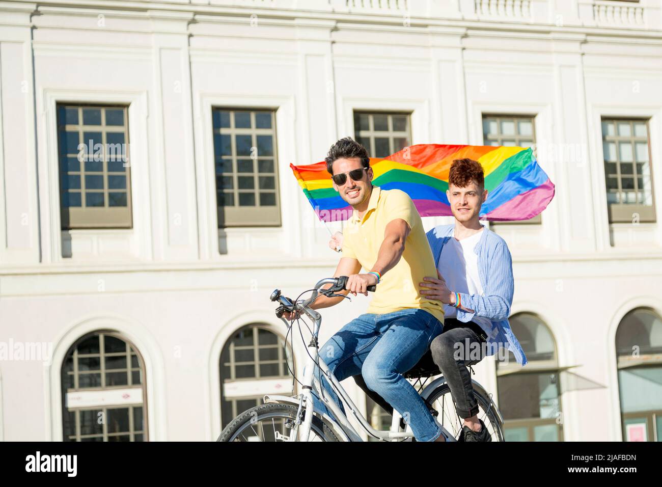 Young gay men couple riding bicycle with gay pride flag looking at camera outdoors. lgbt concept Stock Photo