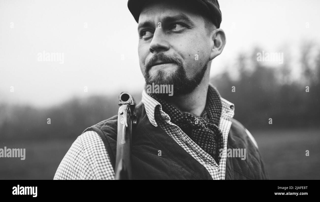 Portrait of hunter man in traditional shooting clothes on field holding shotgun, black and white photo. Stock Photo